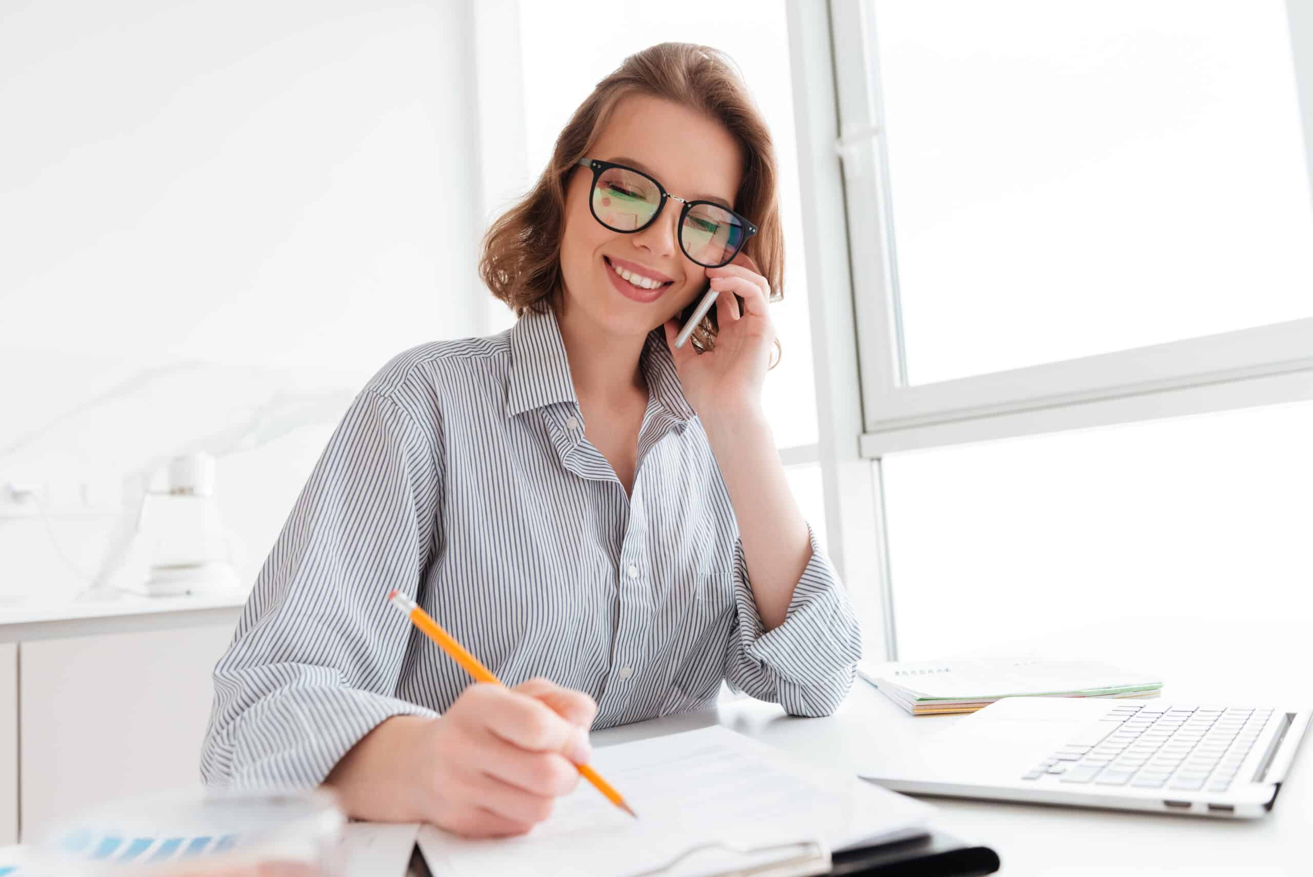 Young assistant in glasses talking on mobile phone while working with documents