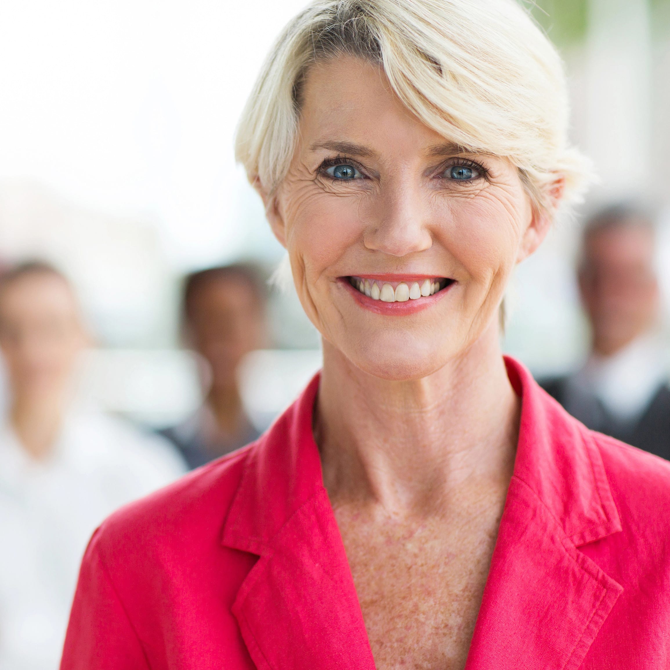 Smiling professional woman with short blonde hair wearing a red blazer, standing confidently in an office setting with coworkers softly blurred in the background.