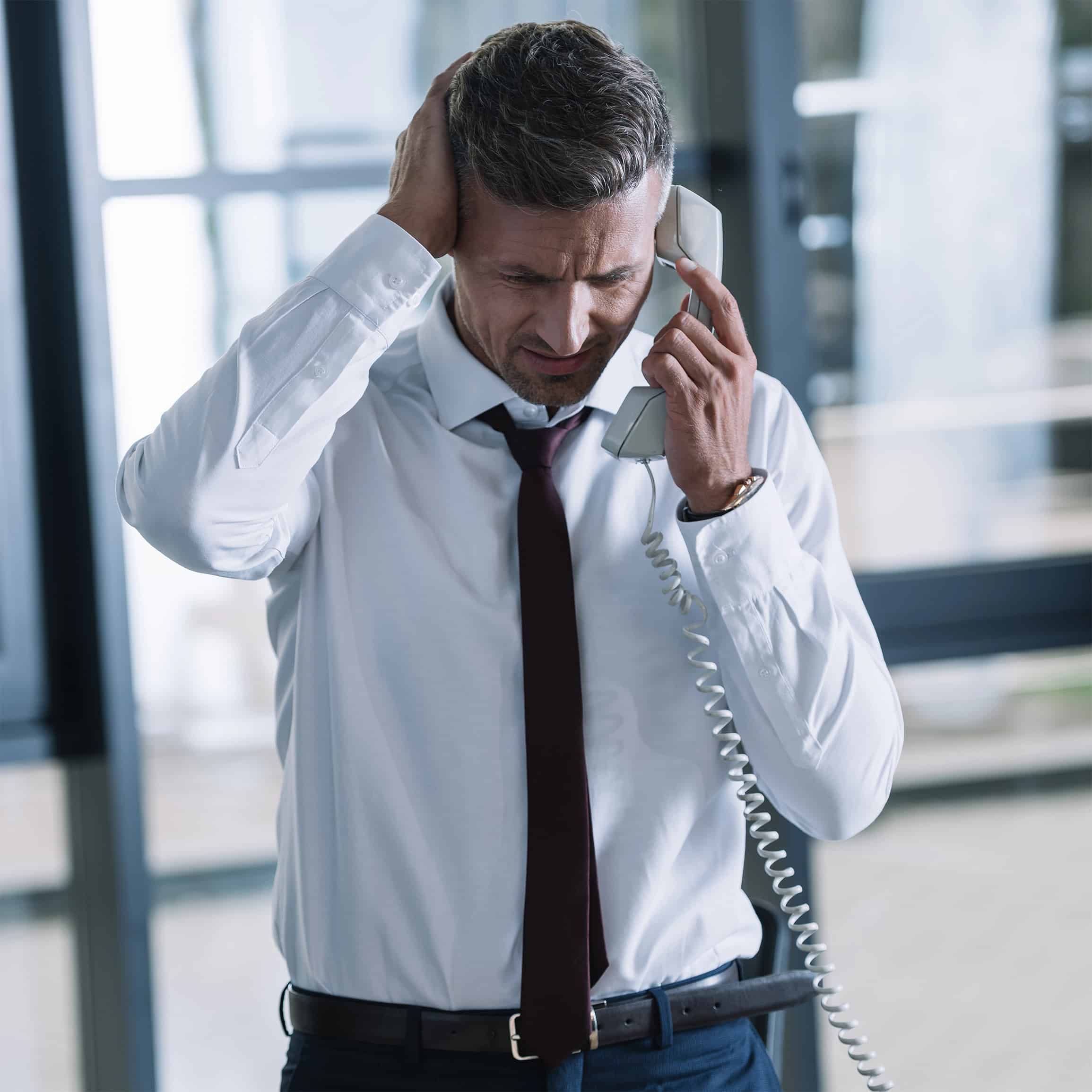 Businessman holds a corded phone and clutches his head, looking stressed during a call.