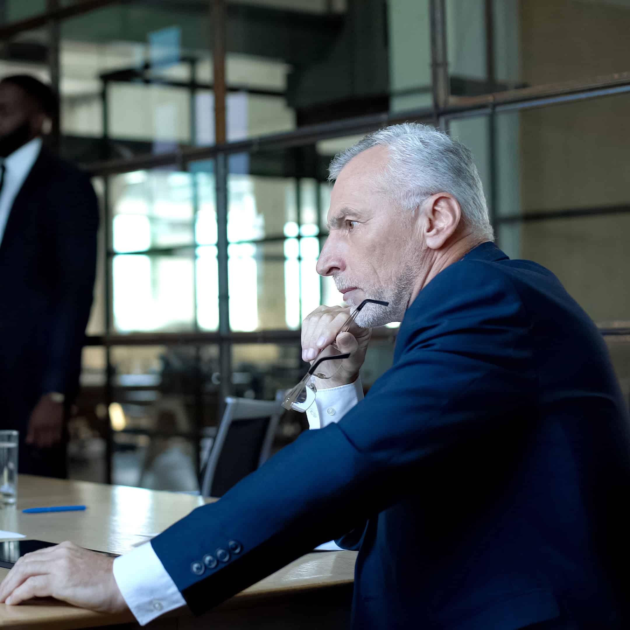 Gray-haired, sullen man in a suit, holding glasses, thinking at a conference table.