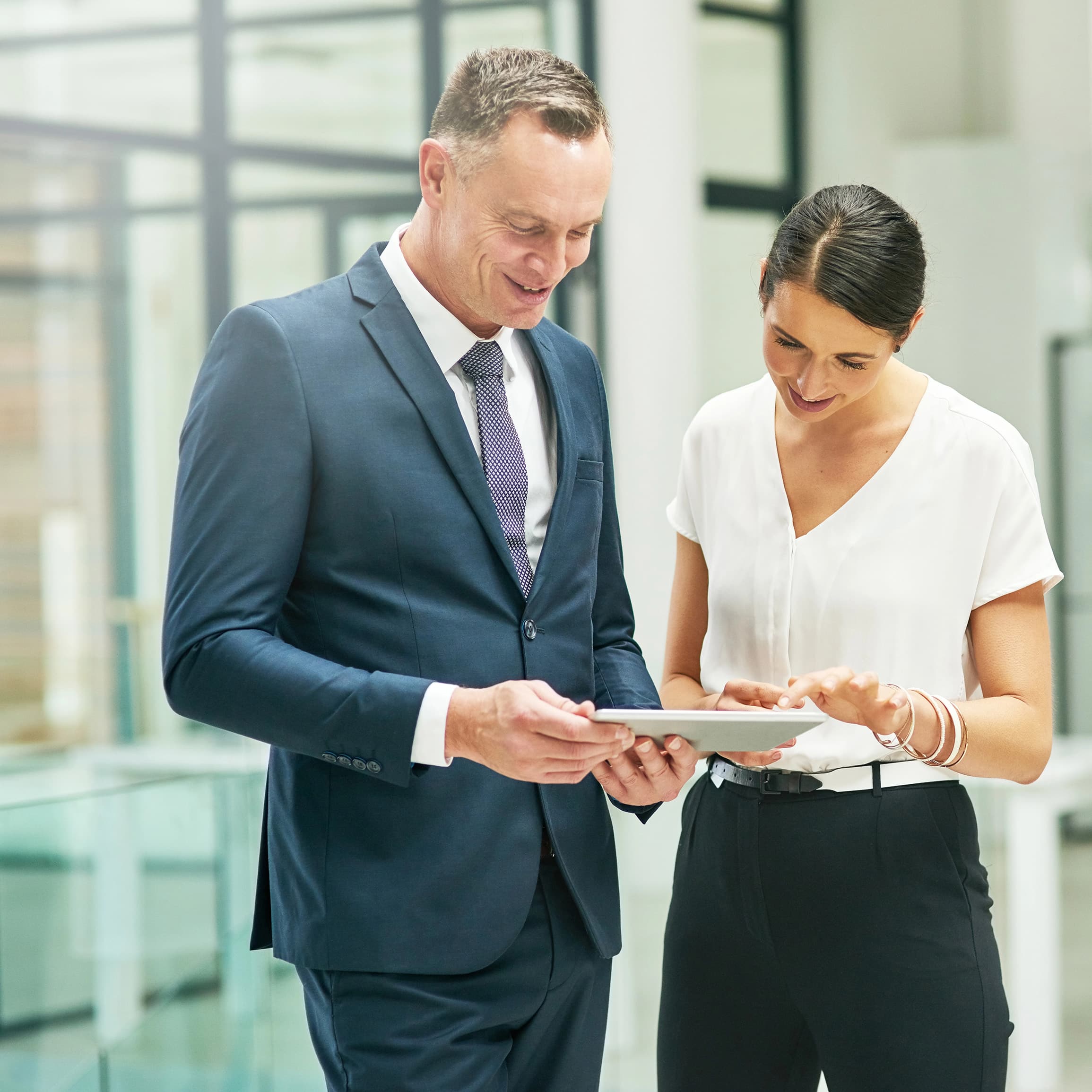 A business man and his Executive Assistant review a tablet together in a bright modern office.