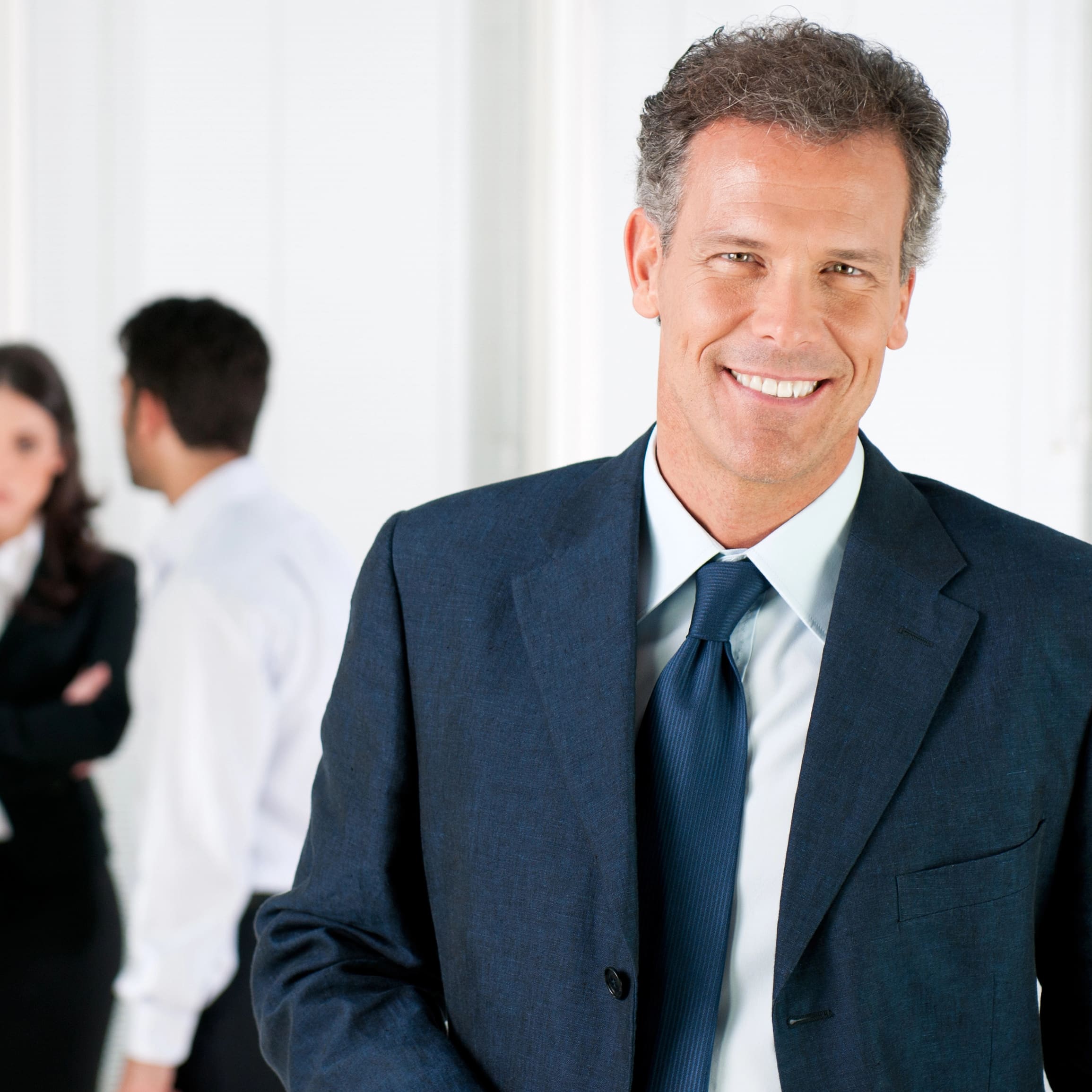 Smiling man in suit in foreground; coworkers converse out of focus behind.